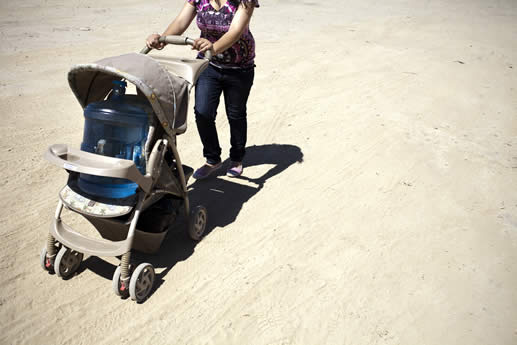 photo of a woman in a dry landscape pushing a baby stroller; in the stroller is a plastic 20 liter jug of water