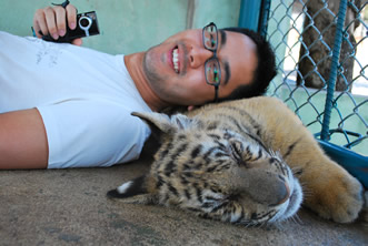 photo of a smiling man holding a mobile phone resting his head on a tiger cub, both are inside a cage