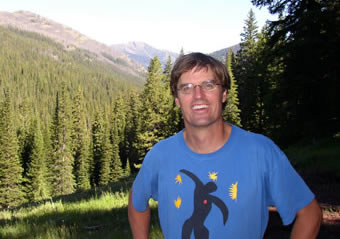 photo of a smiling man in a meadow fringed by pines in sunlight
