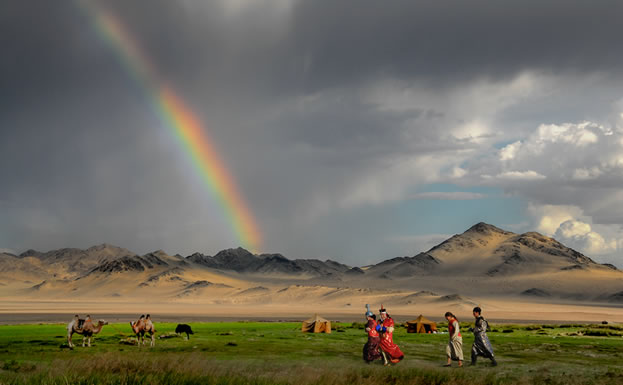 photo of people in the outdoors, with camels and a rainbow