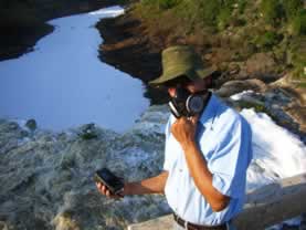photo of a man in a respirator standing on a river beach, he is holding a scientific measuring meter