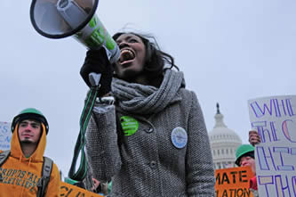 photo of people wearing green hardhats at a demonstration; US Capitol building in the background, young woman with a megaphone in foreground