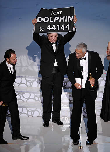 photo of three men in tuxedos, one holding an Oscar, one holding a sign over his head that says: text dolphin to 44144