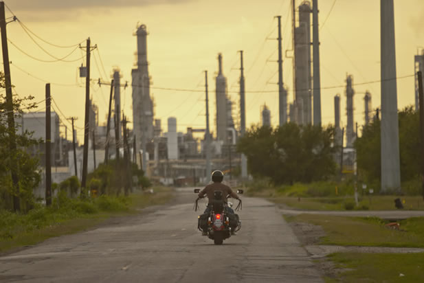 photo of a motorcyclist riding along a curbless street into a cluster of fractionation towers at a refinery. The sky is an unhealthy color.