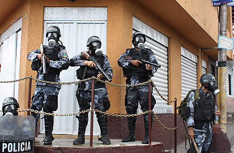 photo of three gas-mask and aromor wearing police, holding tear-gas launchers, word 'policia' on their helmets and shields