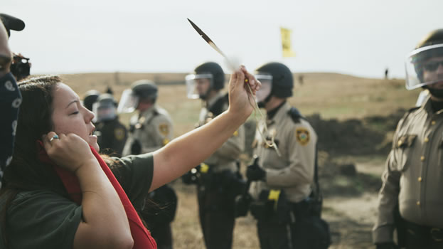 photo of a woman praying, holding a feather to the sky - she's faced with armed and armored law enforcement
